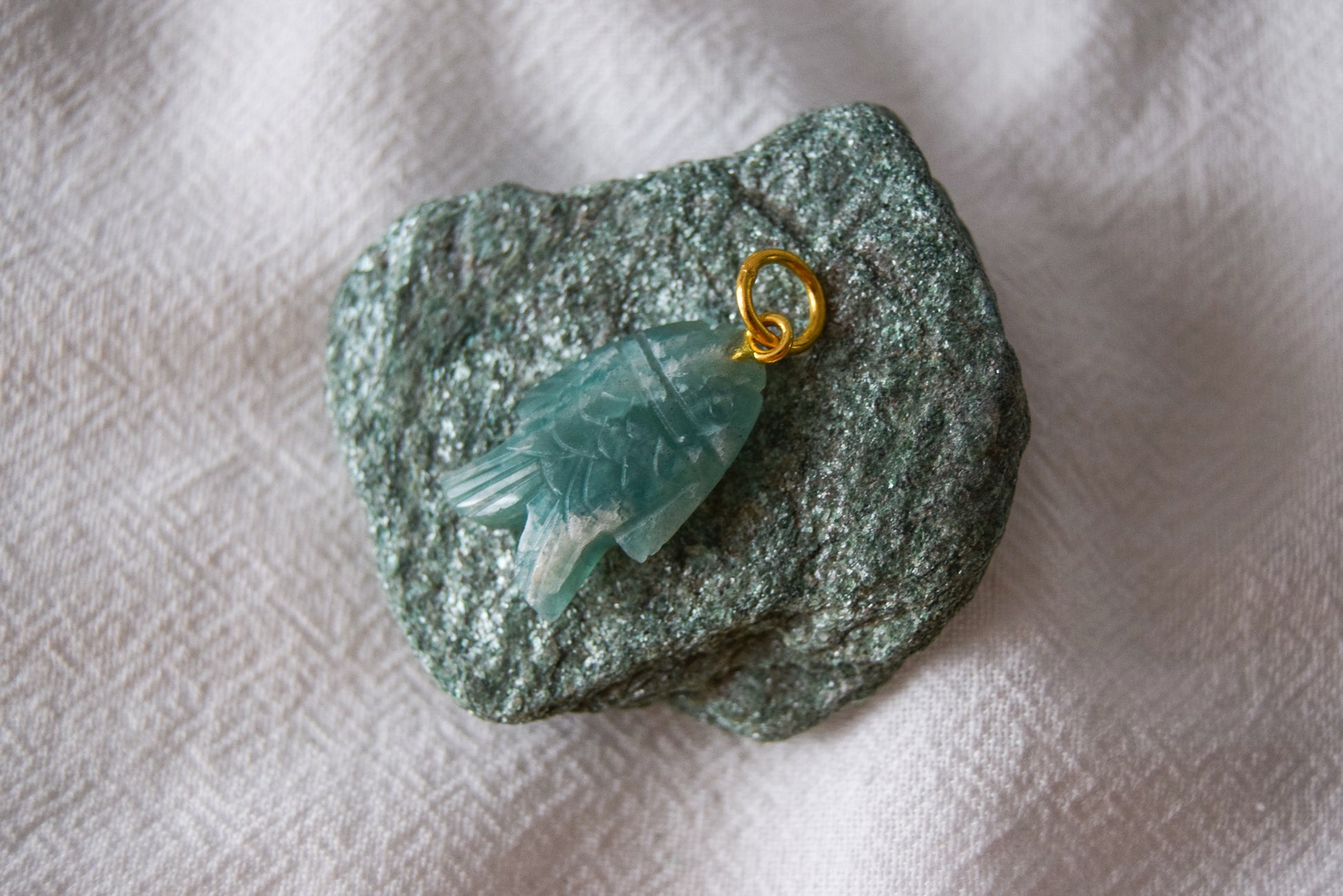 Intricately detailed, this versatile amazonite fish charm features a delicate fish design carved from amazonite gemstone, with the pendant ring made of gold plated material, it is  perfect for various jewelry creations, displayed on a stone and textile background.