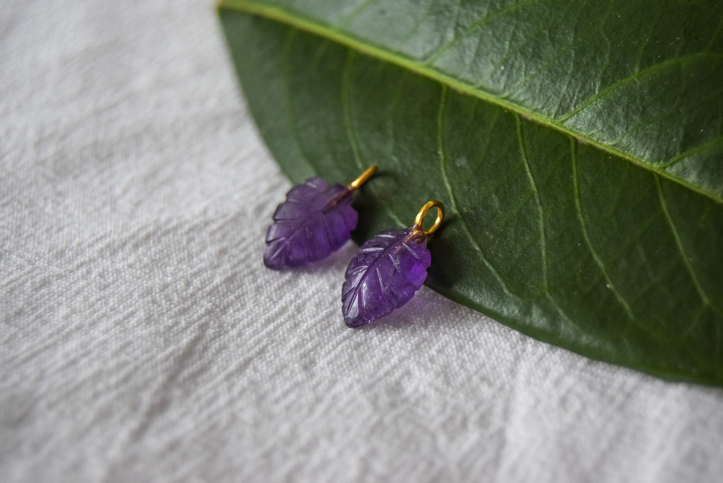 Intricately detailed, this versatile amethyst leaf shaped charm features a delicate leaf design carved from amethyst gemstone, with the pendant ring made of gold plated material, it is  perfect for various jewelry creations like a necklace or earrings, displayed on a leafy background.