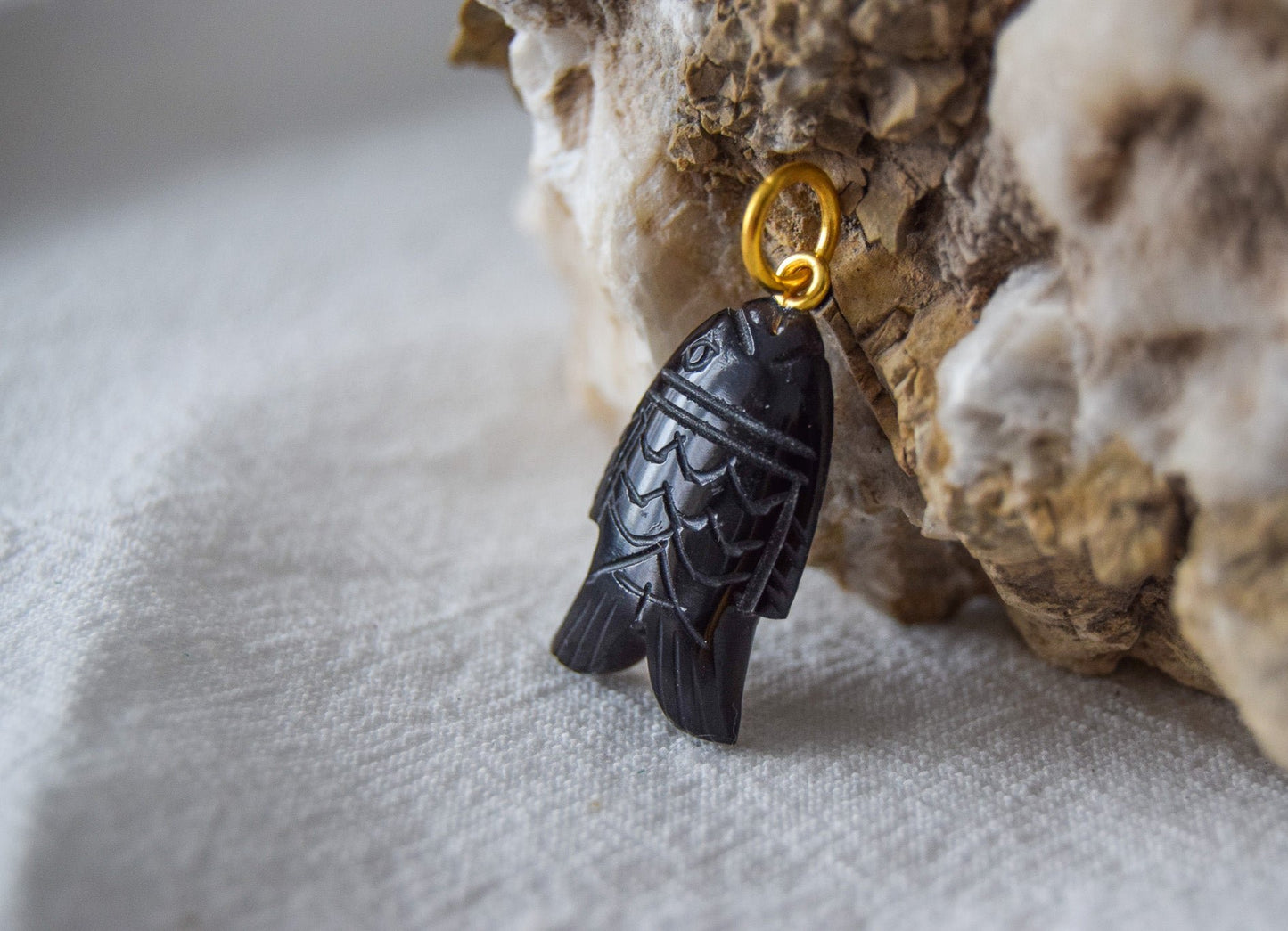 Intricately detailed, this versatile black onyx fish shaped charm features a delicate fish design carved from black onyx gemstone, with the pendant ring made of gold plated material, it is  perfect for various jewelry creations like a necklace or earrings, displayed on a natural stone background.