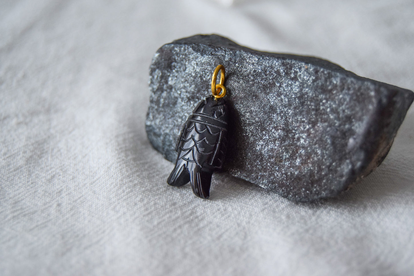 Intricately detailed, this versatile black onyx fish shaped charm features a delicate fish design carved from black onyx gemstone, with the pendant ring made of gold plated material, it is  perfect for various jewelry creations like a necklace or earrings, displayed on a natural black stone and white textile background.