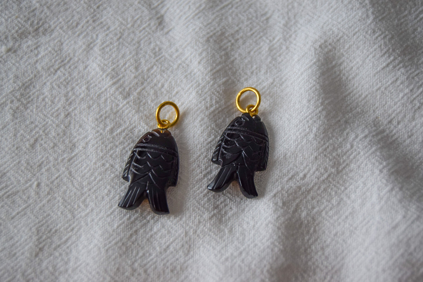 Intricately detailed, this versatile black onyx fish shaped charm features a delicate fish design carved from black onyx gemstone, with the pendant ring made of gold plated material, it is  perfect for various jewelry creations like a necklace or earrings, displayed on a white textile background.