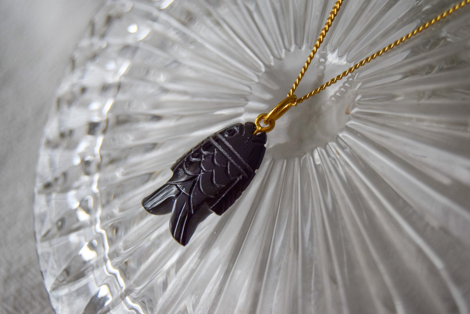 Intricately detailed, this versatile black onyx fish shaped charm features a delicate fish design carved from black onyx gemstone, with the pendant ring made of gold plated material, it is  perfect for various jewelry creations like a necklace or earrings, displayed on a crystal glass background.