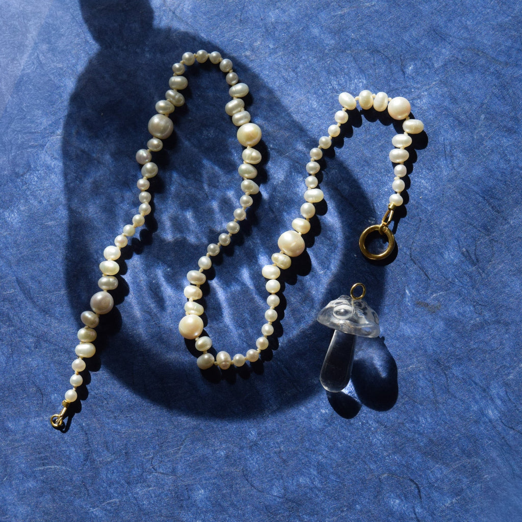 A beaded necklace with irregular cream-colored pearls and gold-toned lock, displayed alongside a clear mushroom-shaped glass charm on a textured blue background with a strong shadow.