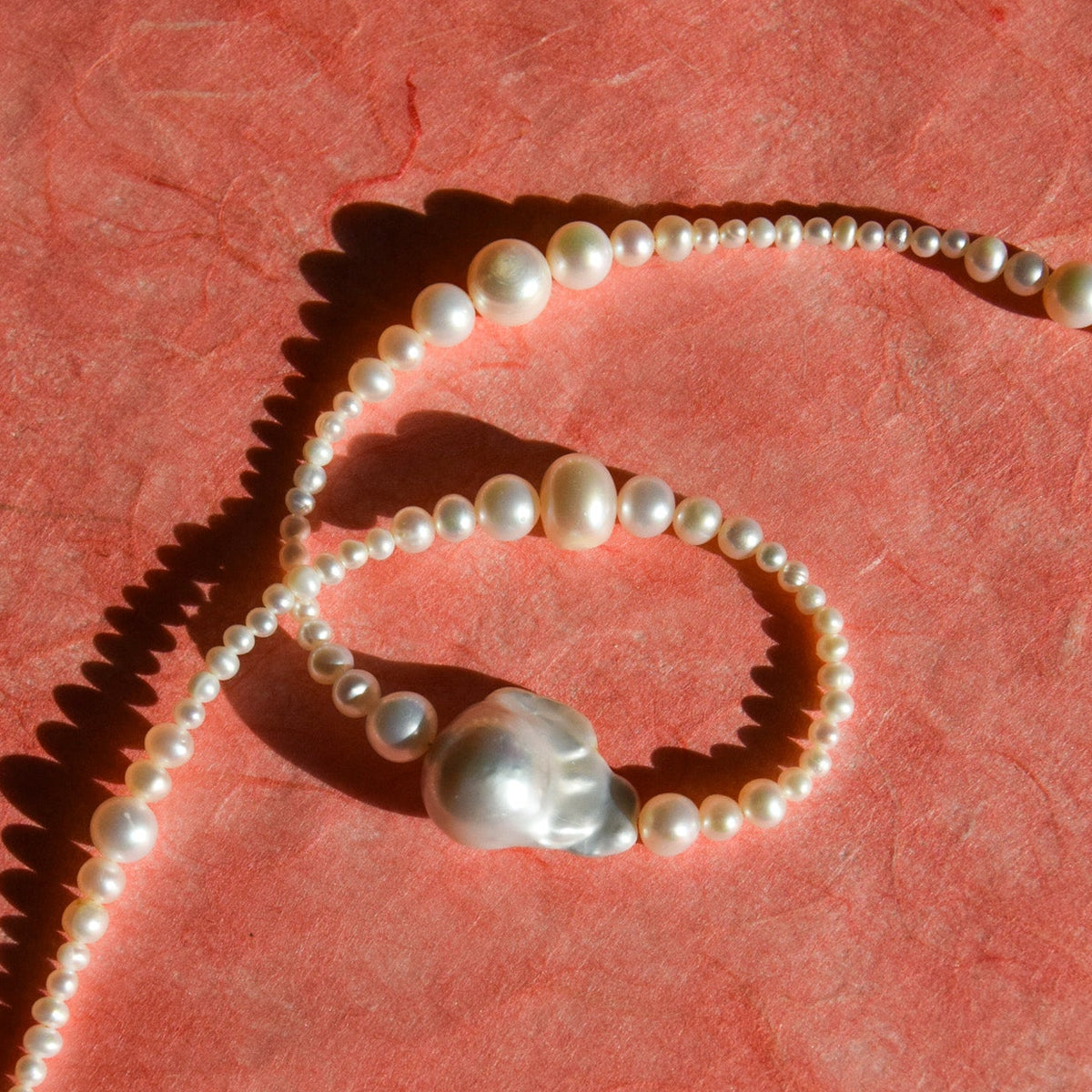 A pearl beaded necklace with a large irregular baroque pearl pendant, styled on a textured pink paper background.