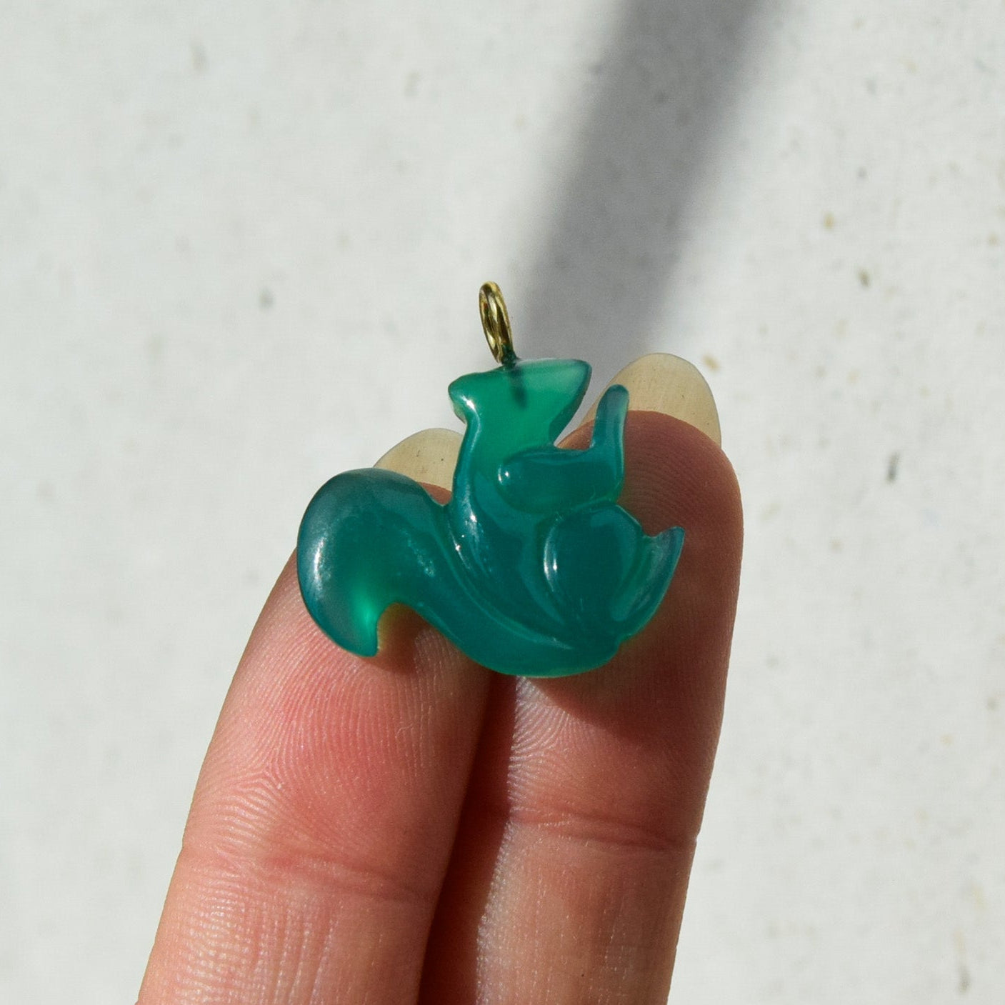 A small carved pendant in a translucent teal green color, held between two fingers to show its scale. The pendant features a stylized, abstract design with a small gold-colored loop at the top.