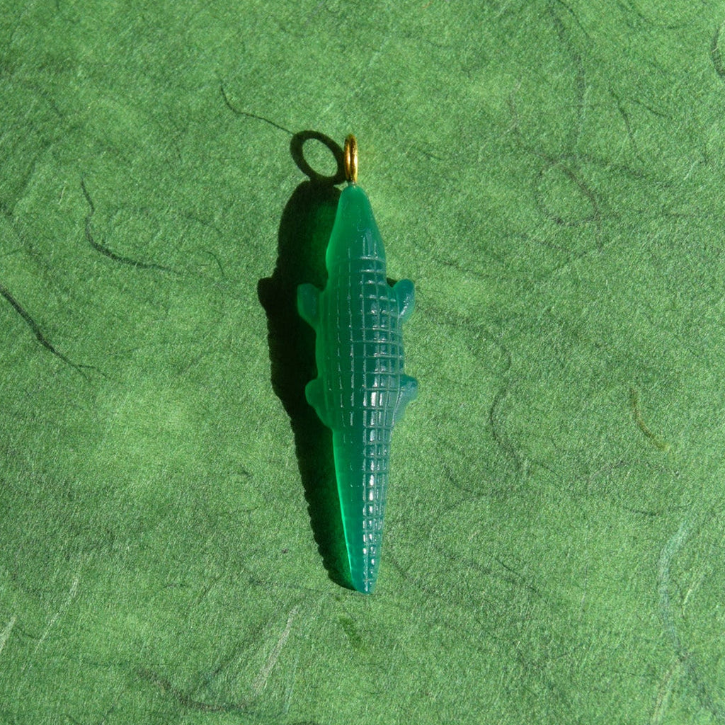 A small green alligator-shaped pendant with a gold-colored loop at the top, resting on a textured green surface.
