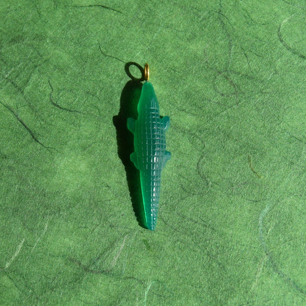 A small green alligator-shaped pendant with a gold-colored loop at the top, resting on a textured green surface.