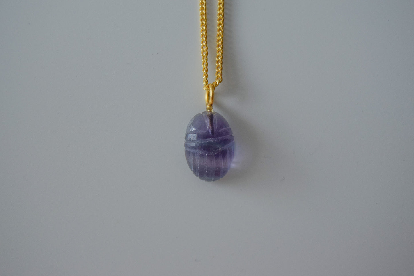 Intricately detailed, this versatile flourite scarab charm features a delicate scarab design carved from flourite gemstone, with the pendant ring made of gold plated material, it is  perfect for various jewelry creations like a necklace or earrings, displayed on a neutral background.
