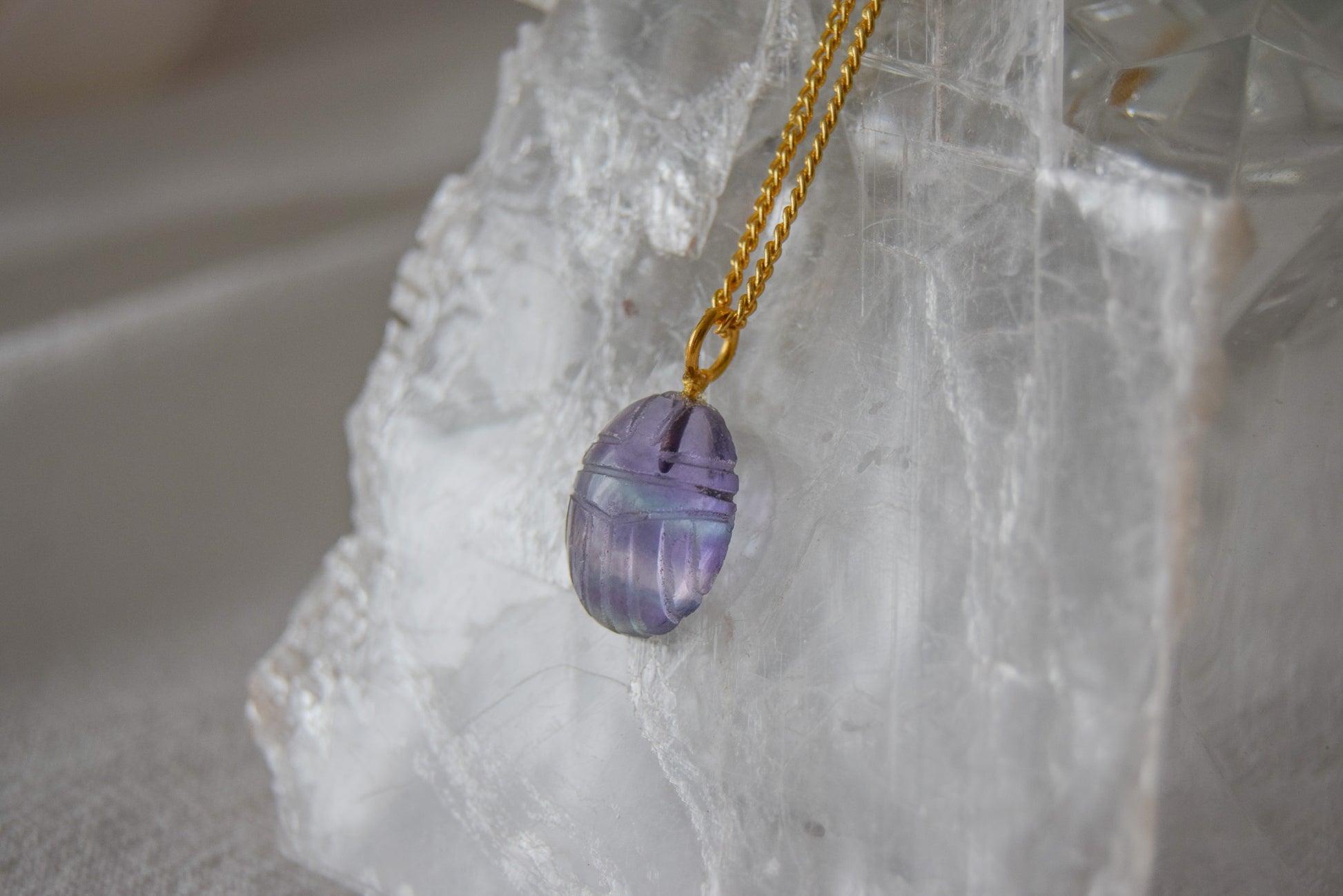 Intricately detailed, this versatile flourite scarab charm features a delicate scarab design carved from flourite gemstone, with the pendant ring made of gold plated material, it is  perfect for various jewelry creations like a necklace or earrings, displayed on a crystal background