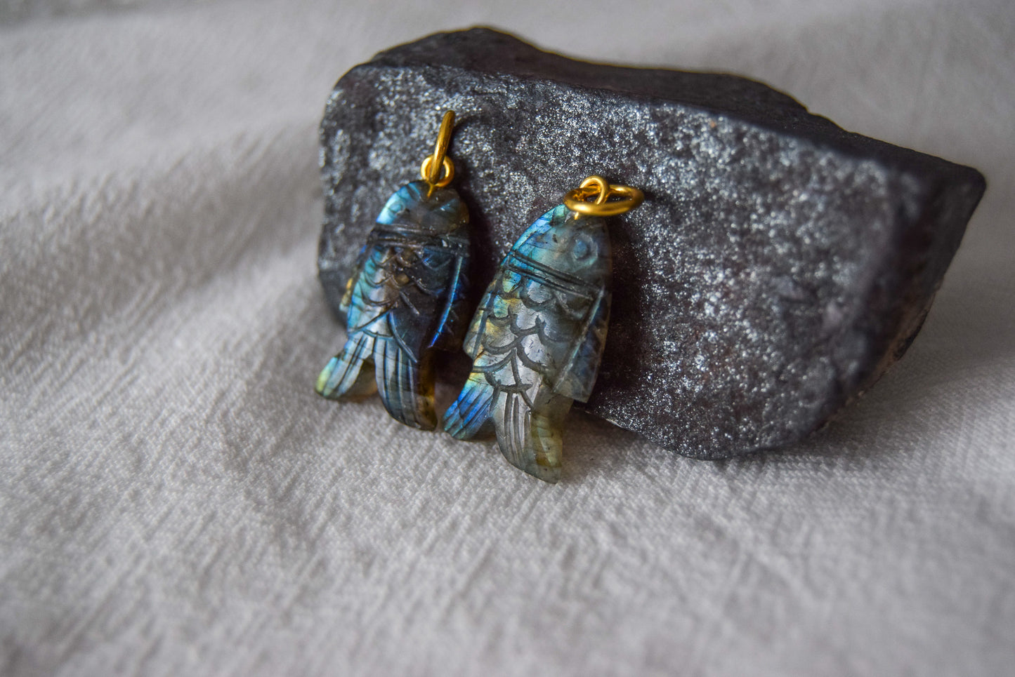 Intricately detailed, this versatile labradorite fish charm features a delicate fish design carved from labradorite gemstone, with the pendant ring made of gold plated material, it is  perfect for various jewelry creations like a necklace or earrings, displayed on a natural black stone and white textile background