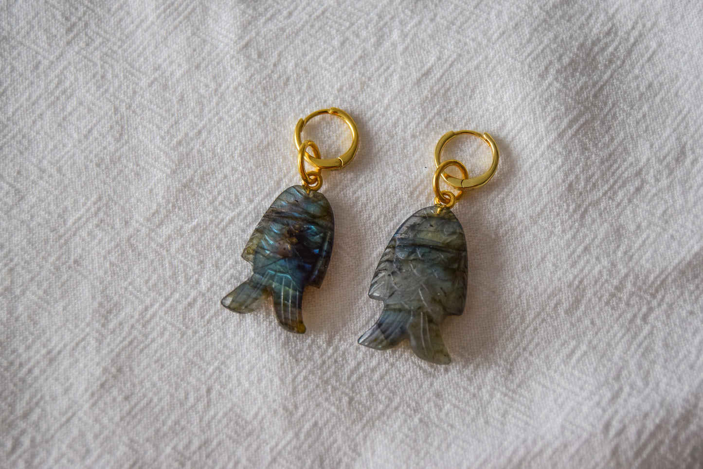 Intricately detailed, this versatile labradorite fish charm features a delicate fish design carved from labradorite gemstone, with the pendant ring made of gold plated material, it is  perfect for various jewelry creations like a necklace or earrings, displayed on a white textile background