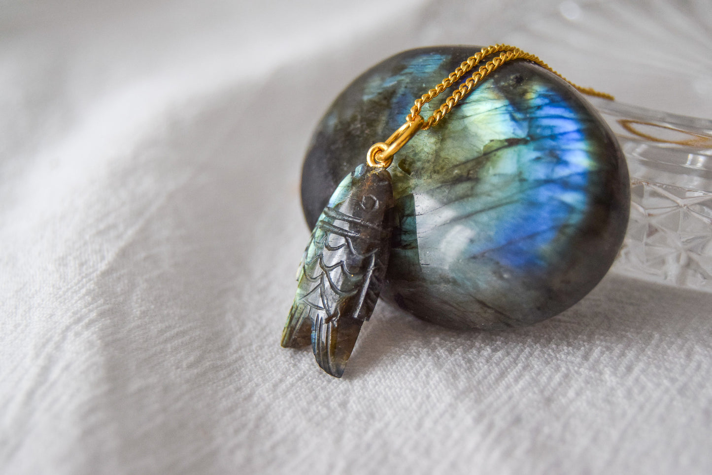 Intricately detailed, this versatile labradorite fish charm features a delicate fish design carved from labradorite gemstone, with the pendant ring made of gold plated material, it is  perfect for various jewelry creations like a necklace or earrings, displayed on a labradorite and crystal glass background