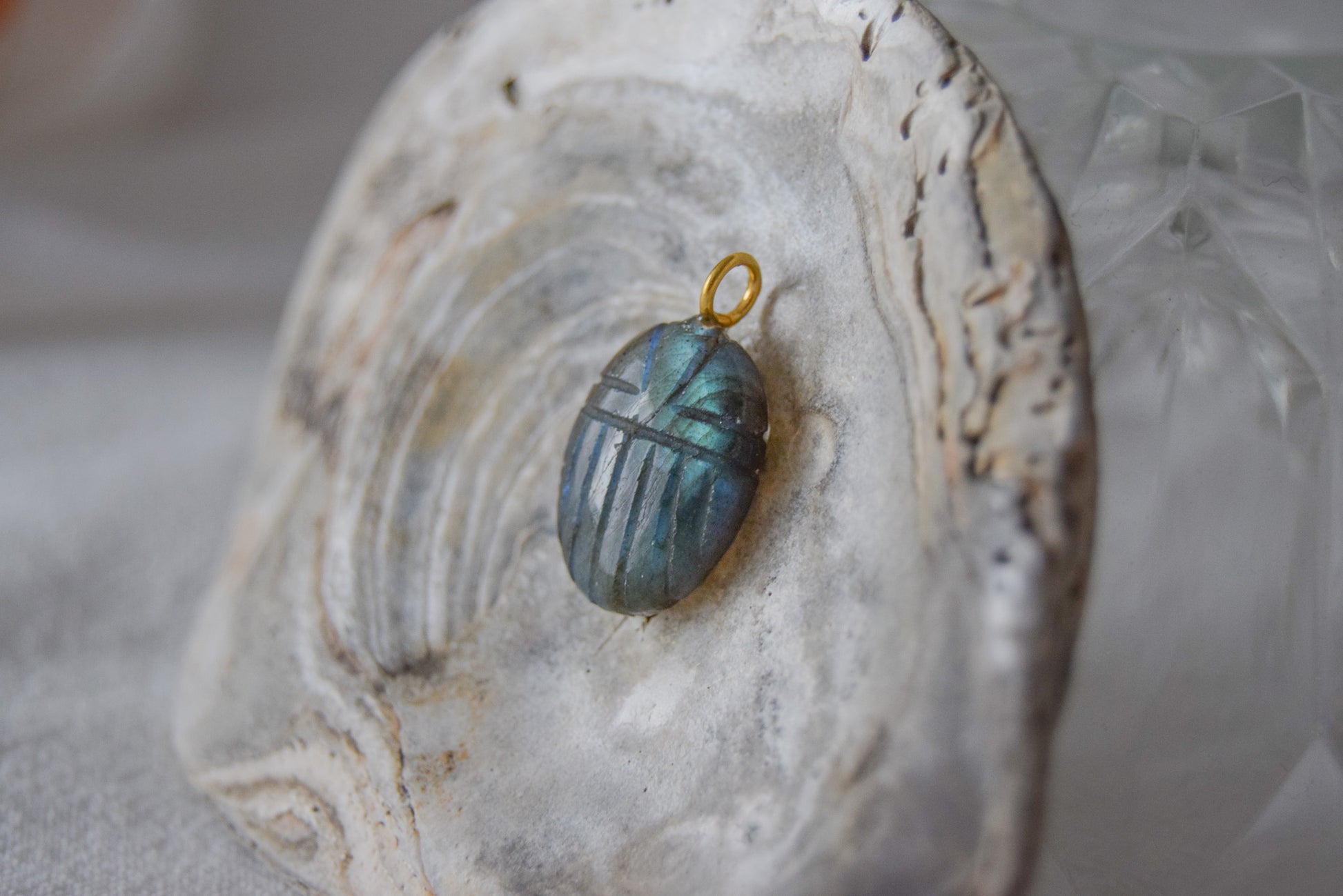 Intricately detailed, this versatile labradorite scarab charm features a delicate scarab design carved from labradorite gemstone, with the pendant ring made of gold plated material, it is  perfect for various jewelry creations like a necklace or earrings, displayed on a shell background