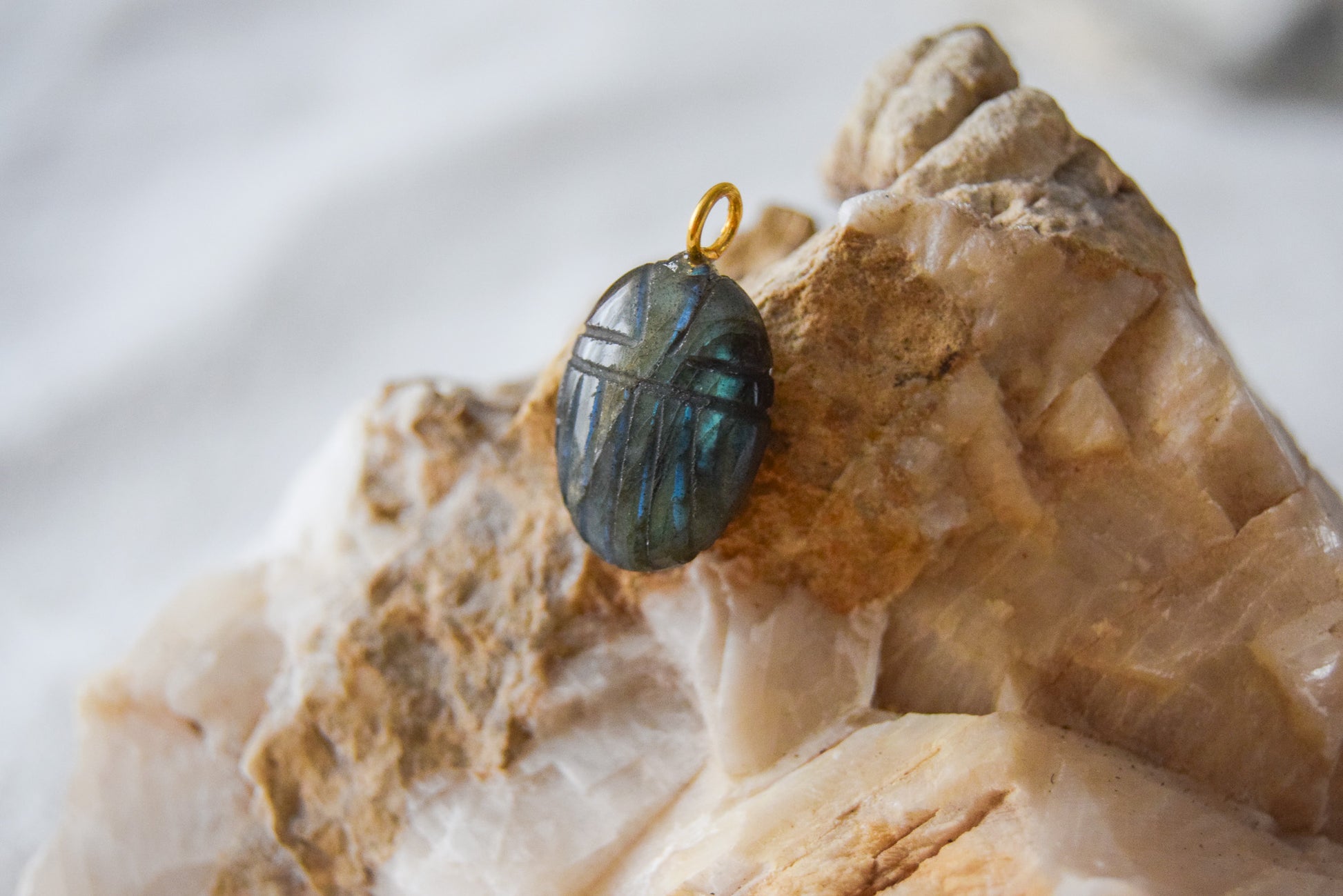 Intricately detailed, this versatile labradorite scarab charm features a delicate scarab design carved from labradorite gemstone, with the pendant ring made of gold plated material, it is  perfect for various jewelry creations like a necklace or earrings, displayed on a natural stone background