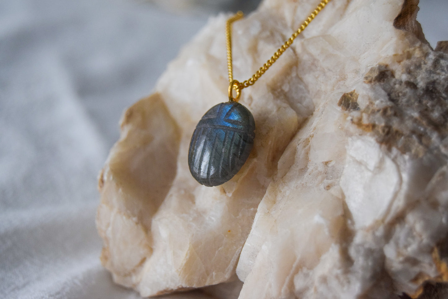 Intricately detailed, this versatile labradorite scarab charm features a delicate scarab design carved from labradorite gemstone, with the pendant ring made of gold plated material, it is  perfect for various jewelry creations like a necklace or earrings, displayed on a natural stone background