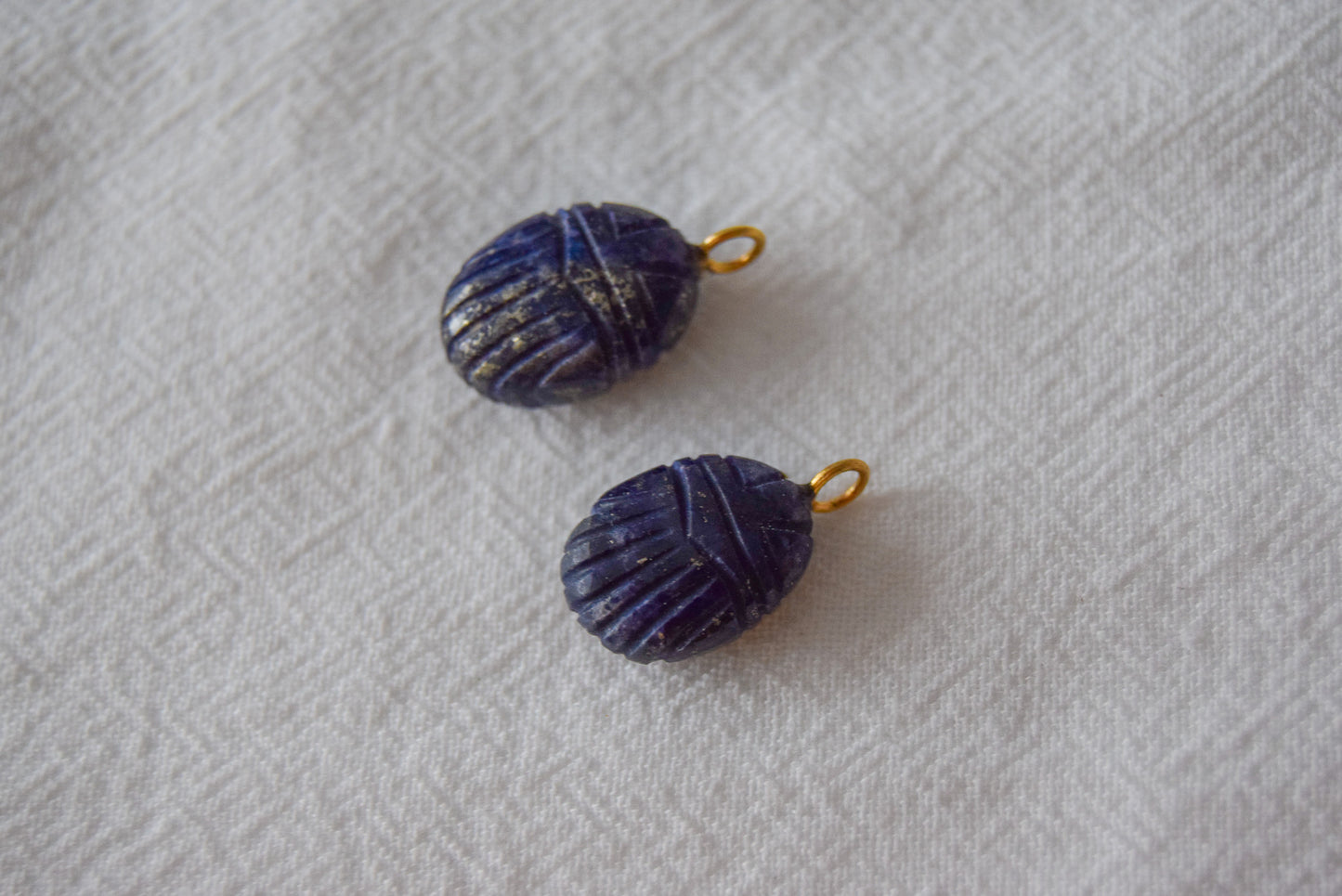 Intricately detailed, this versatile lapis lazuli scarab charm features a delicate scarab design carved from lapis lazuli gemstone, with the pendant ring made of gold plated material, it is  perfect for various jewelry creations like a necklace or earrings, displayed on a white textile background
