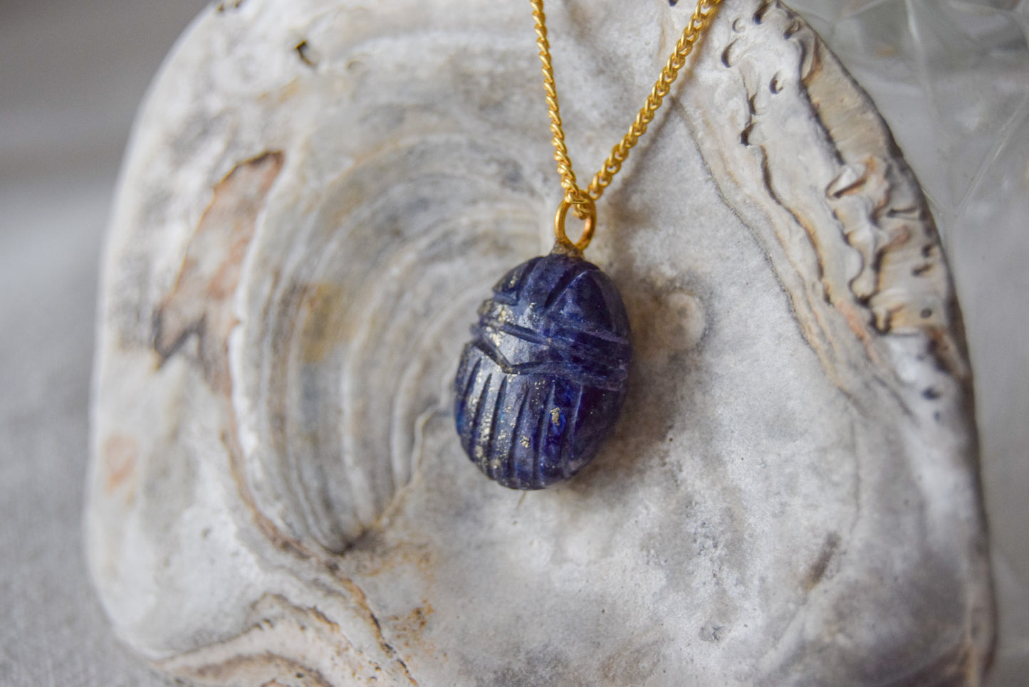 Intricately detailed, this versatile lapis lazuli scarab charm features a delicate scarab design carved from lapis lazuli gemstone, with the pendant ring made of gold plated material, it is  perfect for various jewelry creations like a necklace or earrings, displayed on a shell background