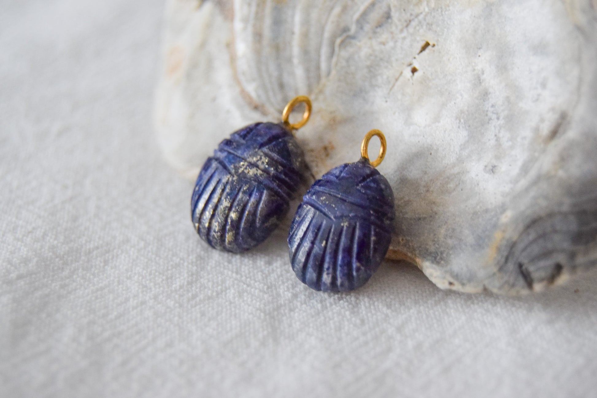 Intricately detailed, this versatile lapis lazuli scarab charm features a delicate scarab design carved from lapis lazuli gemstone, with the pendant ring made of gold plated material, it is  perfect for various jewelry creations like a necklace or earrings, displayed on a shell background