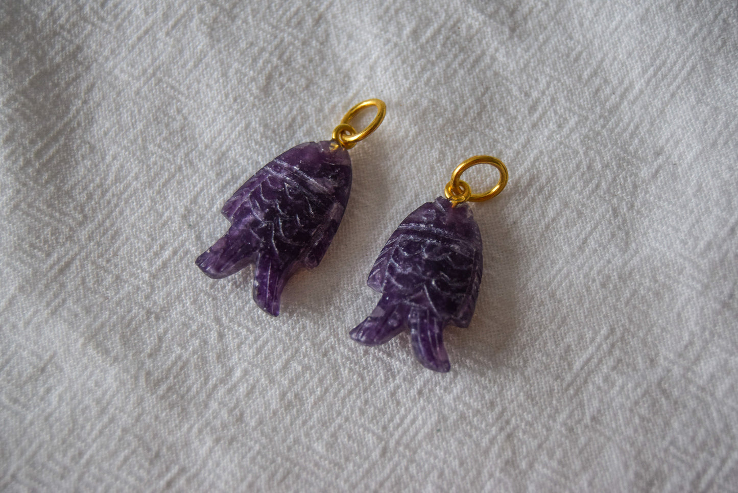 Intricately detailed, this versatile lepidolite fish charm features a delicate fish design carved from lepidolite gemstone, with the pendant ring made of gold plated material, it is perfect for various jewelry creations like a necklace or earrings, displayed on a white textile background