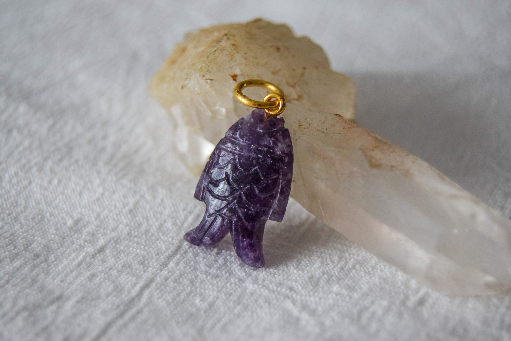 Intricately detailed, this versatile lepidolite fish charm features a delicate fish design carved from lepidolite gemstone, with the pendant ring made of gold plated material, it is perfect for various jewelry creations like a necklace or earrings, displayed on a clear quartz crystal background