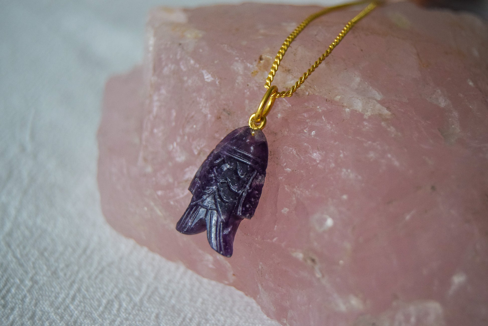 Intricately detailed, this versatile lepidolite fish charm features a delicate fish design carved from lepidolite gemstone, with the pendant ring made of gold plated material, it is perfect for various jewelry creations like a necklace or earrings, displayed on a rose stone background