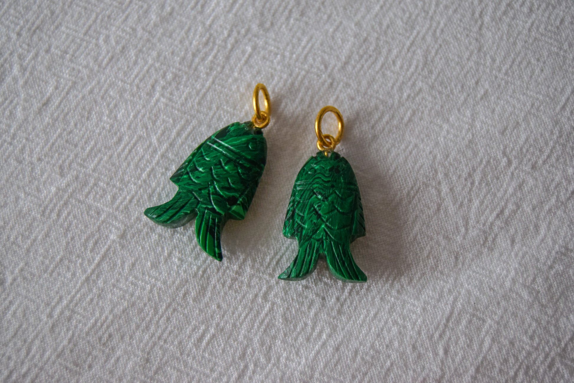 Intricately detailed, this versatile malachite fish charm features a delicate fish design carved from malachite gemstone, with the pendant ring made of gold plated material, it is perfect for various jewelry creations like a necklace or earrings, displayed on a white textile background