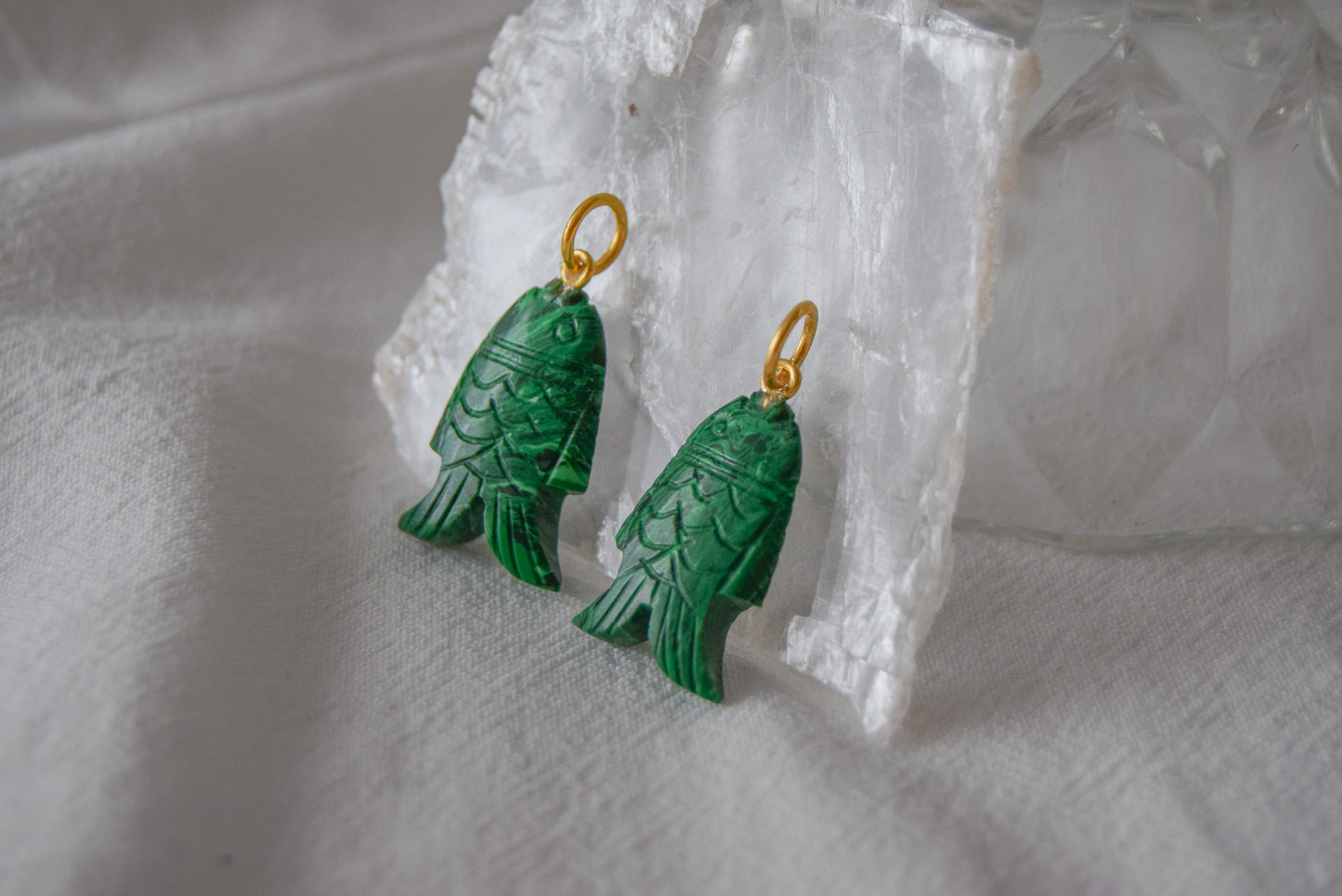 Intricately detailed, this versatile malachite fish charm features a delicate fish design carved from malachite gemstone, with the pendant ring made of gold plated material, it is perfect for various jewelry creations like a necklace or earrings, displayed on a white textile and clear quartz crystal background