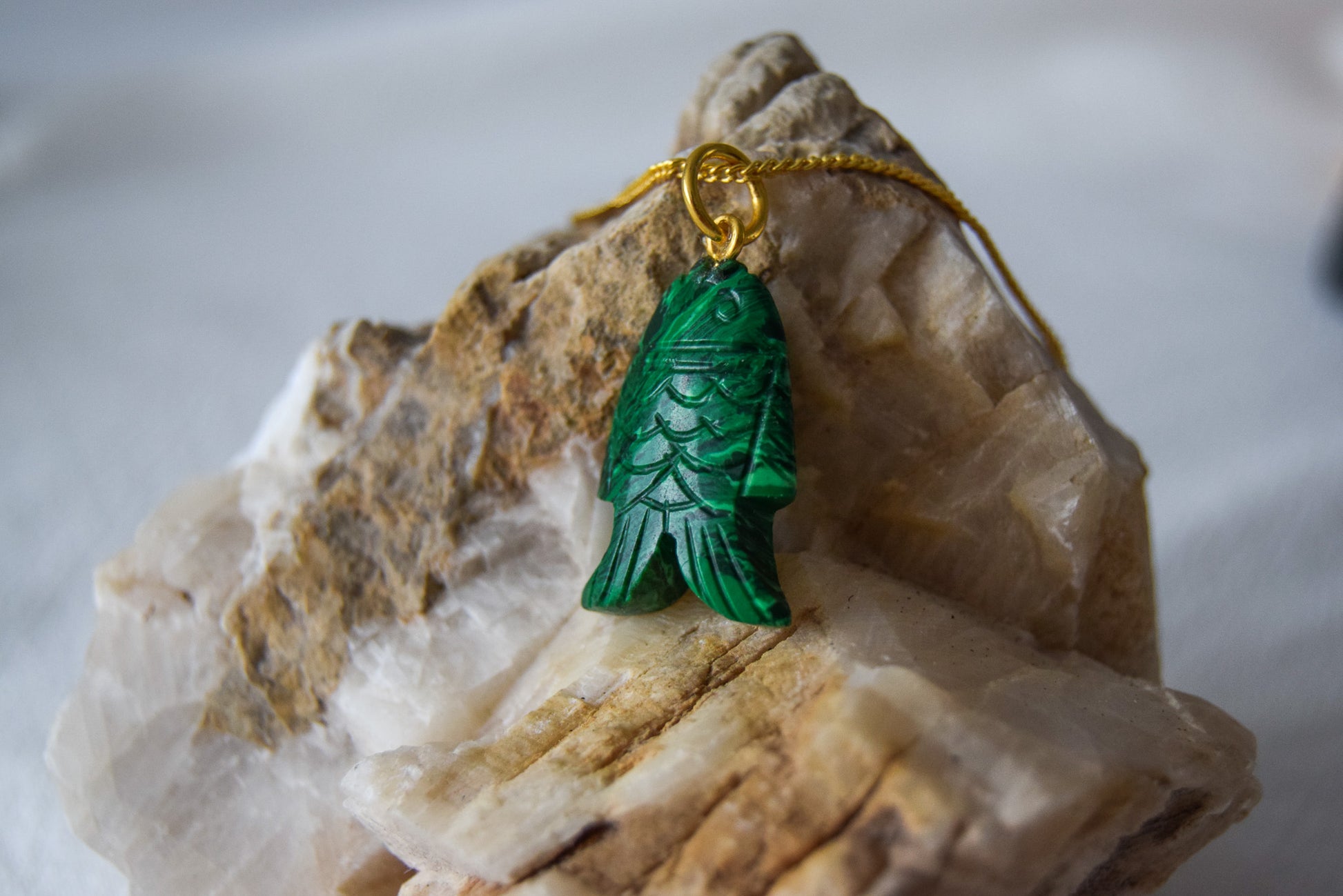 Intricately detailed, this versatile malachite fish charm features a delicate fish design carved from malachite gemstone, with the pendant ring made of gold plated material, it is perfect for various jewelry creations like a necklace or earrings, displayed on a white textile and natural stone background