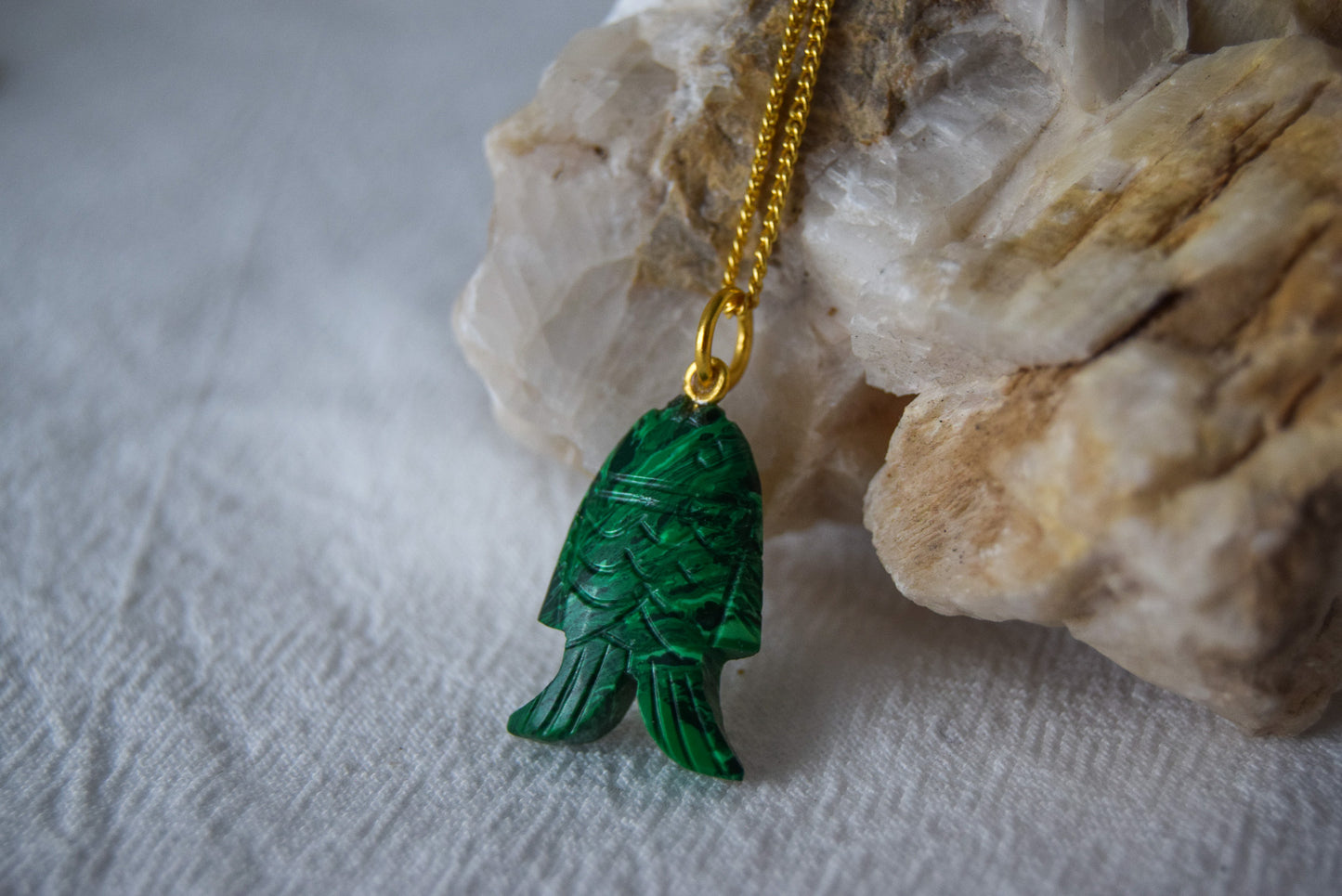 Intricately detailed, this versatile malachite fish charm features a delicate fish design carved from malachite gemstone, with the pendant ring made of gold plated material, it is perfect for various jewelry creations like a necklace or earrings, displayed on a white textile and natural stone background
