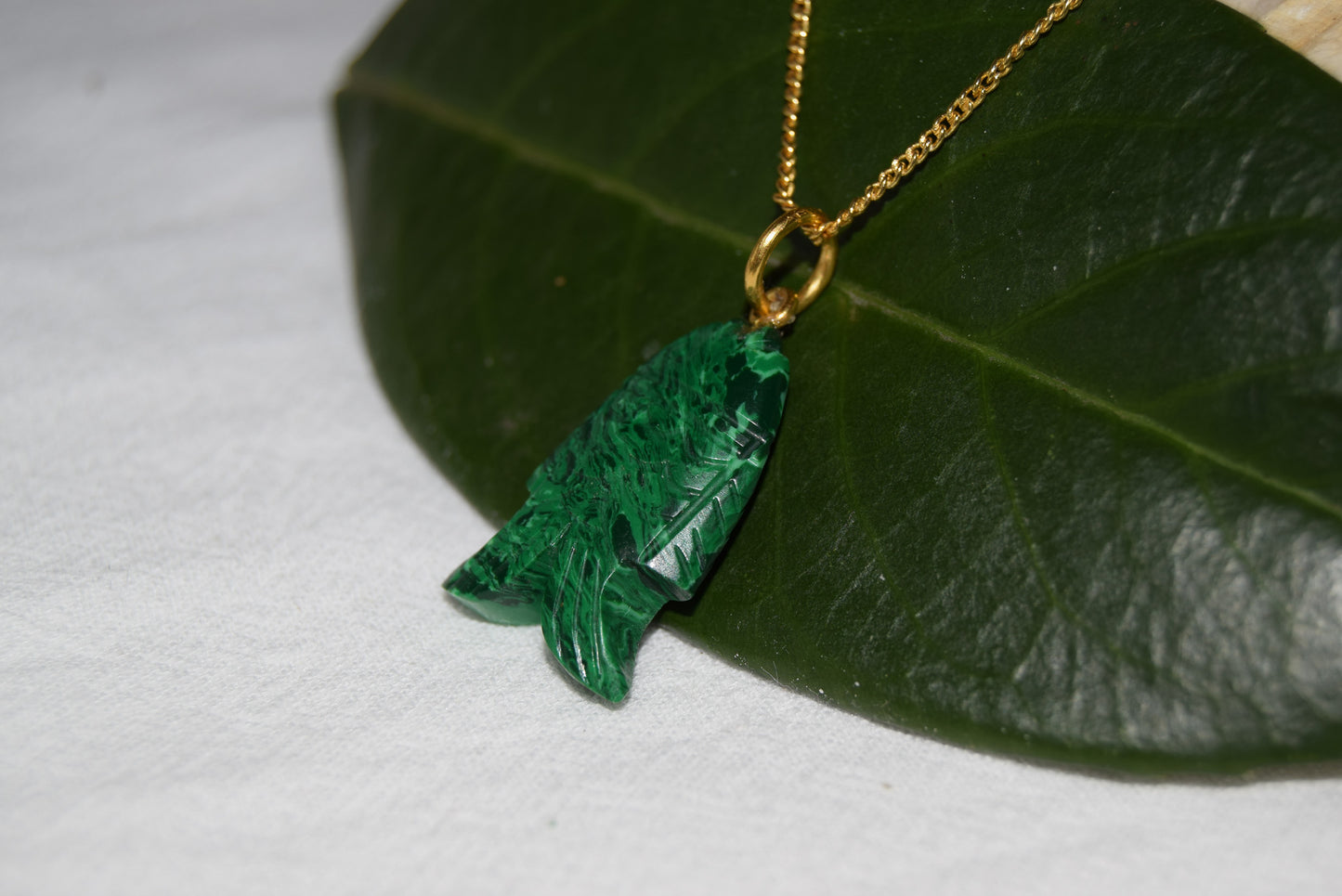 Intricately detailed, this versatile malachite fish charm features a delicate fish design carved from malachite gemstone, with the pendant ring made of gold plated material, it is perfect for various jewelry creations like a necklace or earrings, displayed on a leaf background