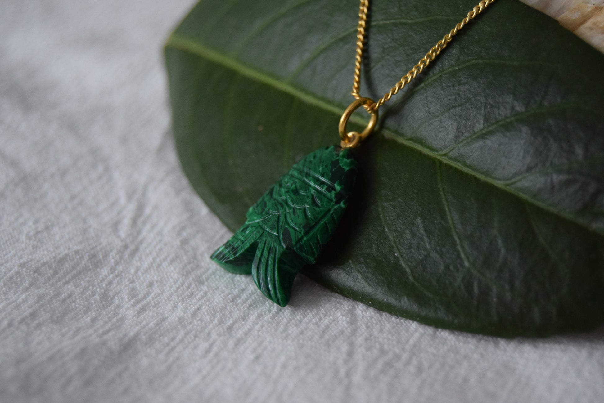 Intricately detailed, this versatile malachite fish charm features a delicate fish design carved from malachite gemstone, with the pendant ring made of gold plated material, it is perfect for various jewelry creations like a necklace or earrings, displayed on a leaf background