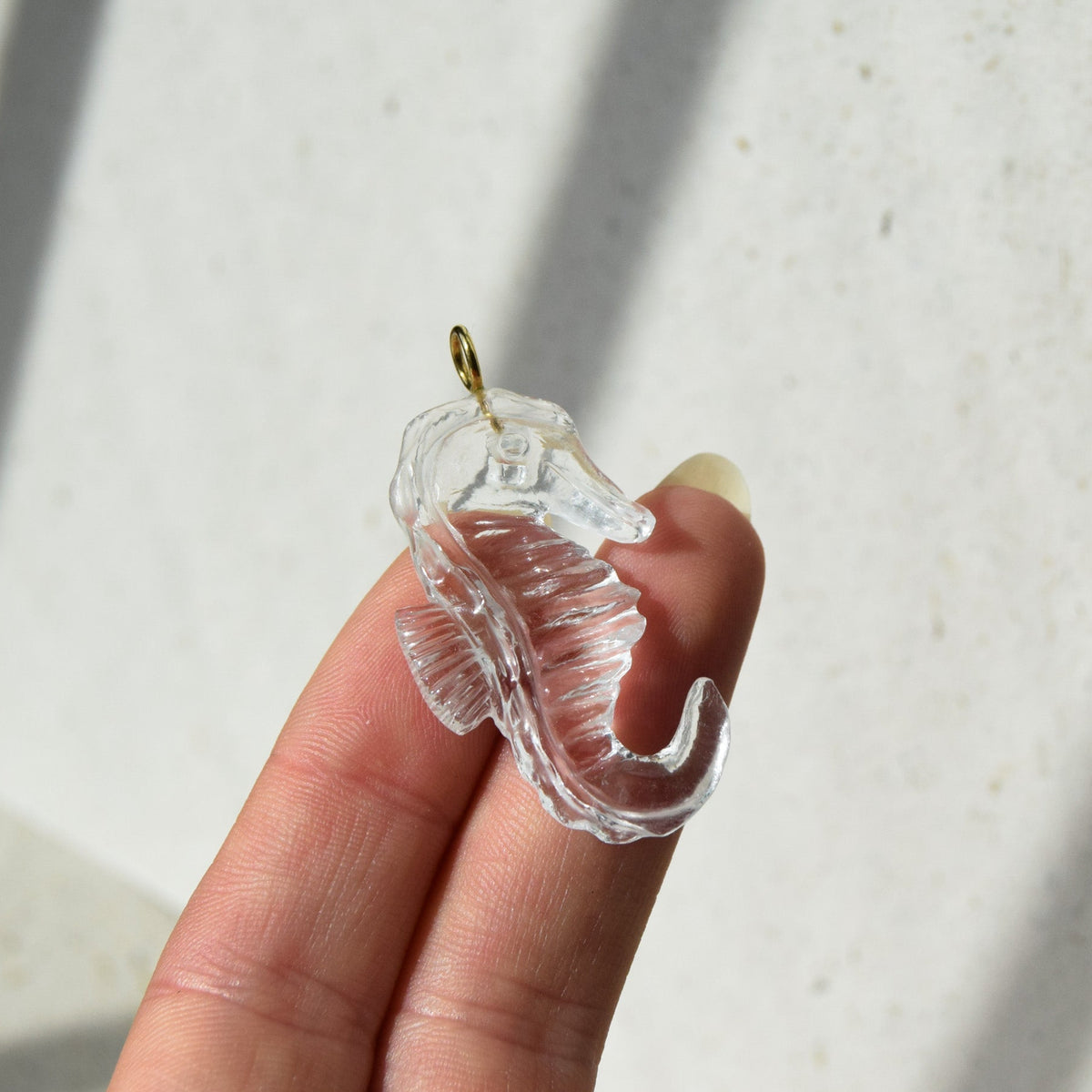 A clear quartz seahorse pendant with a small gold-colored loop at the top, held between a person's fingers against a light off-white background.