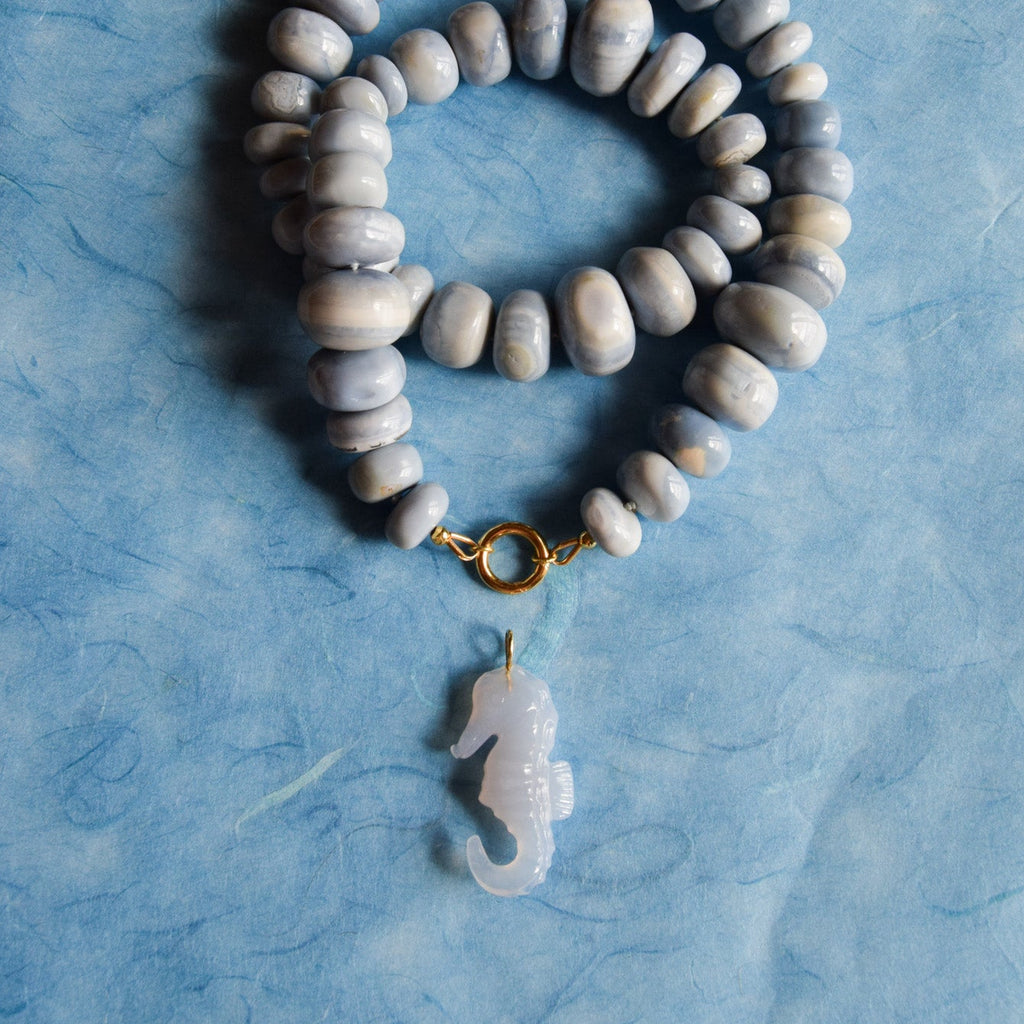 A beaded necklace with smooth, rondelle blue opal beads, a gold toggle clasp, and a translucent blue seahorse pendant, displayed on a textured light blue background.
