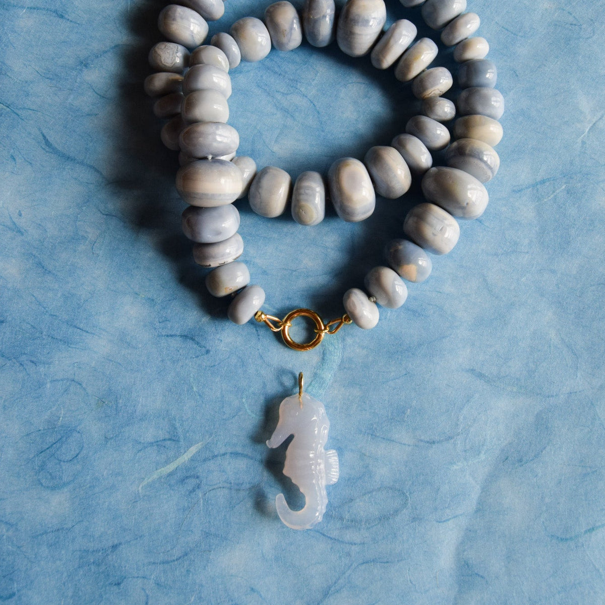 A beaded necklace with smooth, rondelle blue opal beads, a gold toggle clasp, and a translucent blue seahorse pendant, displayed on a textured light blue background.
